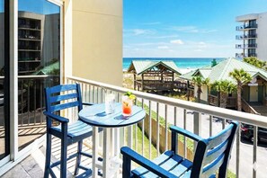 Ein Balkon mit zwei blauen Stühlen und einem kleinen Tisch, der Blick auf einen Strand mit einer Holzkonstruktion und Palmen gerichtet ist.