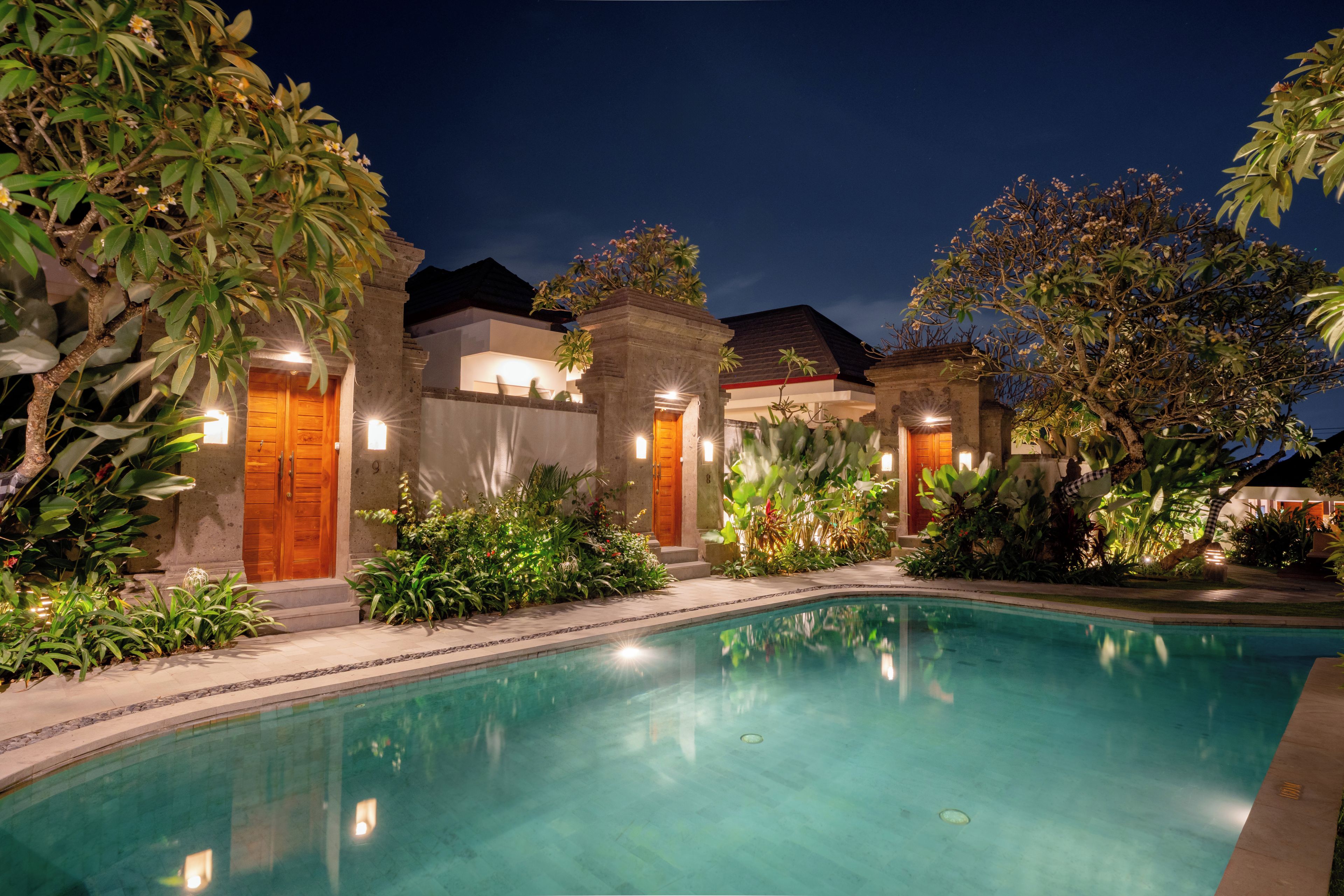 A pool area with a clear pool, surrounded by lush greenery and a building with multiple doors.