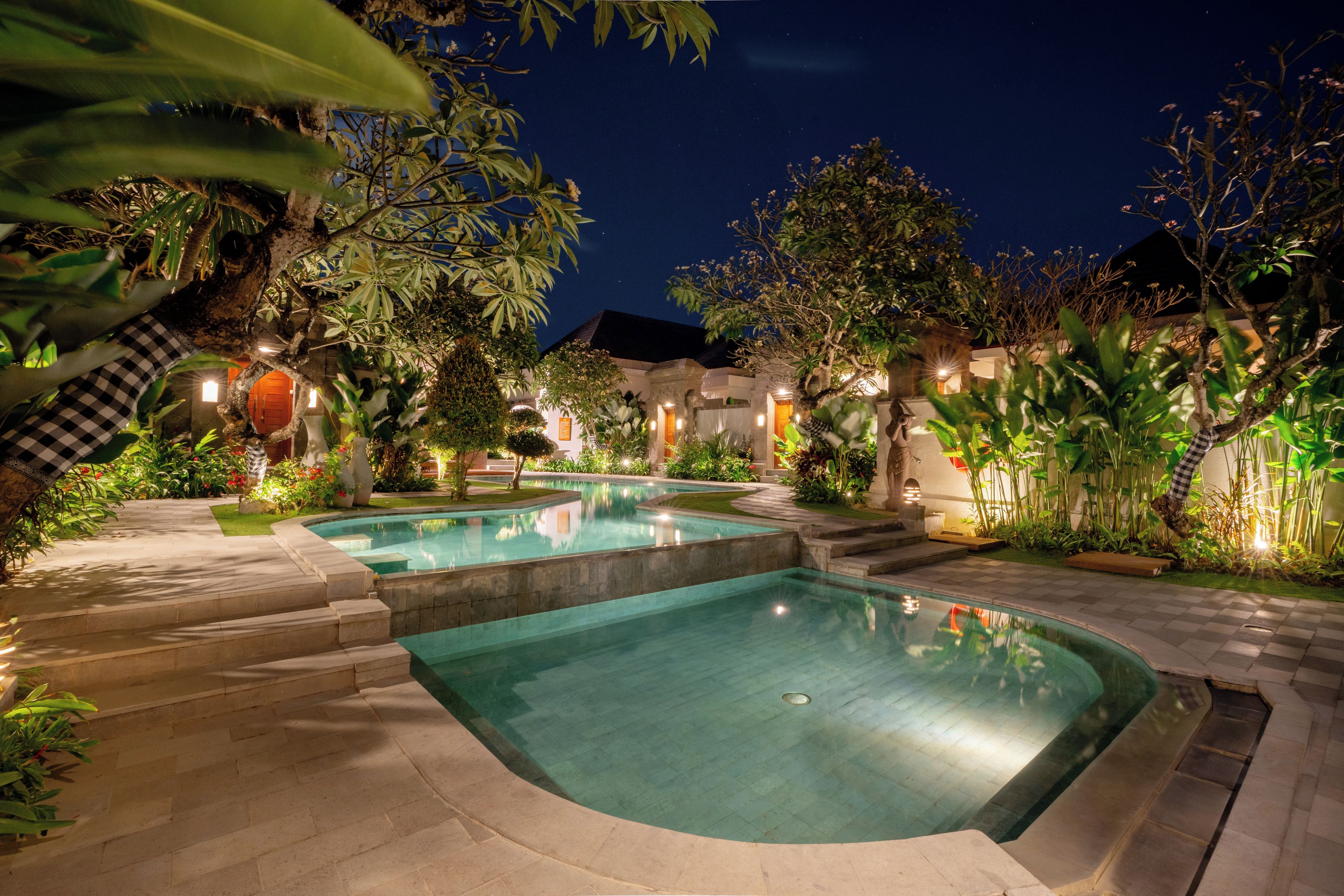 A nighttime view of a swimming pool area with two pools, surrounded by lush greenery and illuminated by ambient lighting.