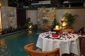 A poolside dining area with a table set for two, surrounded by stone walls and potted plants. - The Prime Seminyak (Kerobokan)