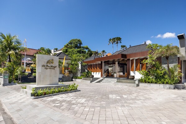 A hotel entrance with a sign, paved walkway, and surrounding greenery. - The Prime Seminyak (Kerobokan)