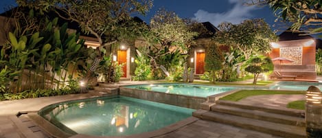 A well-lit outdoor swimming pool area with multiple pools, surrounded by lush greenery and traditional architecture.