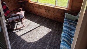 A cozy cabin interior with a rocking chair, a window with a view of a grassy yard, and a sign indicating the cabin's coordinates.