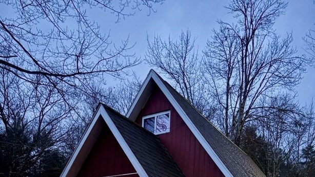 A red wooden cabin with a gabled roof, a large window, and a small attached shed.