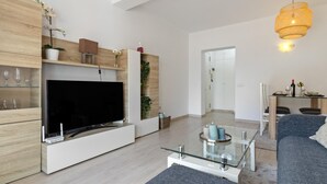 En moderne stue med et fladskærms-tv, træhylder, et glasbord og et spiseområde med bord og stole.