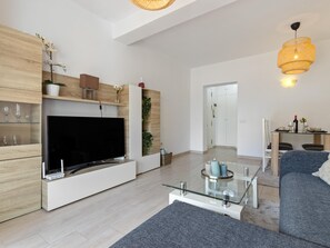 A modern living room with a flat-screen TV, wooden shelving, a glass coffee table, and a dining area with a table and chairs. - Sol Montemar 19 - Terrace, Swimming Pool, Parking, 2 bedrooms (Torremolinos)