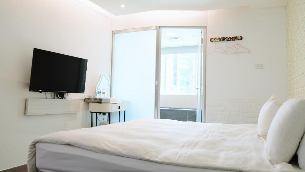 A hotel room with a bed, a flat-screen TV mounted on the wall, a small bedside table, and a sliding glass door leading to a balcony.