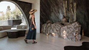 A modern interior with a large arched window, a reception desk with a cactus sculpture, and a seating area with sofas. - One of One - Adults Only (Santorini)