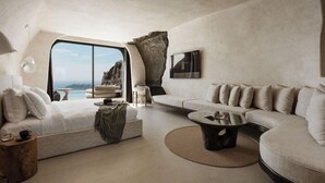 A modern interior with a large window, a sofa, a coffee table, and a unique rock feature. - One of One - Adults Only (Santorini)