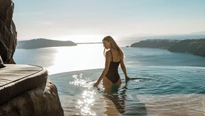 A woman in a swimsuit standing in a clear water body with a view of distant landforms. - One of One - Adults Only (Santorini)