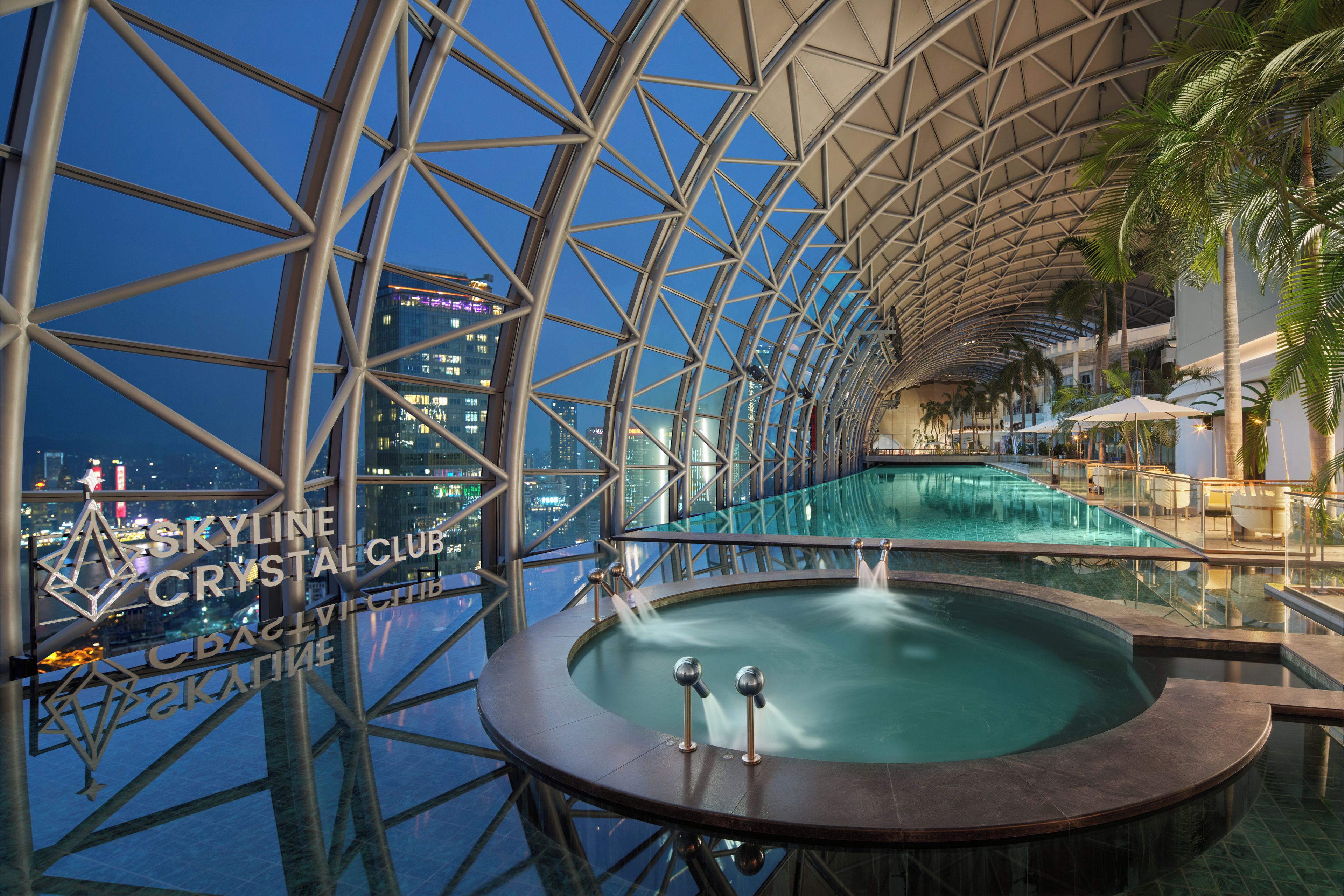 Un área de piscina interior moderna con piso de vidrio, rodeada por una estructura metálica, y vista panorámica de la ciudad de noche.