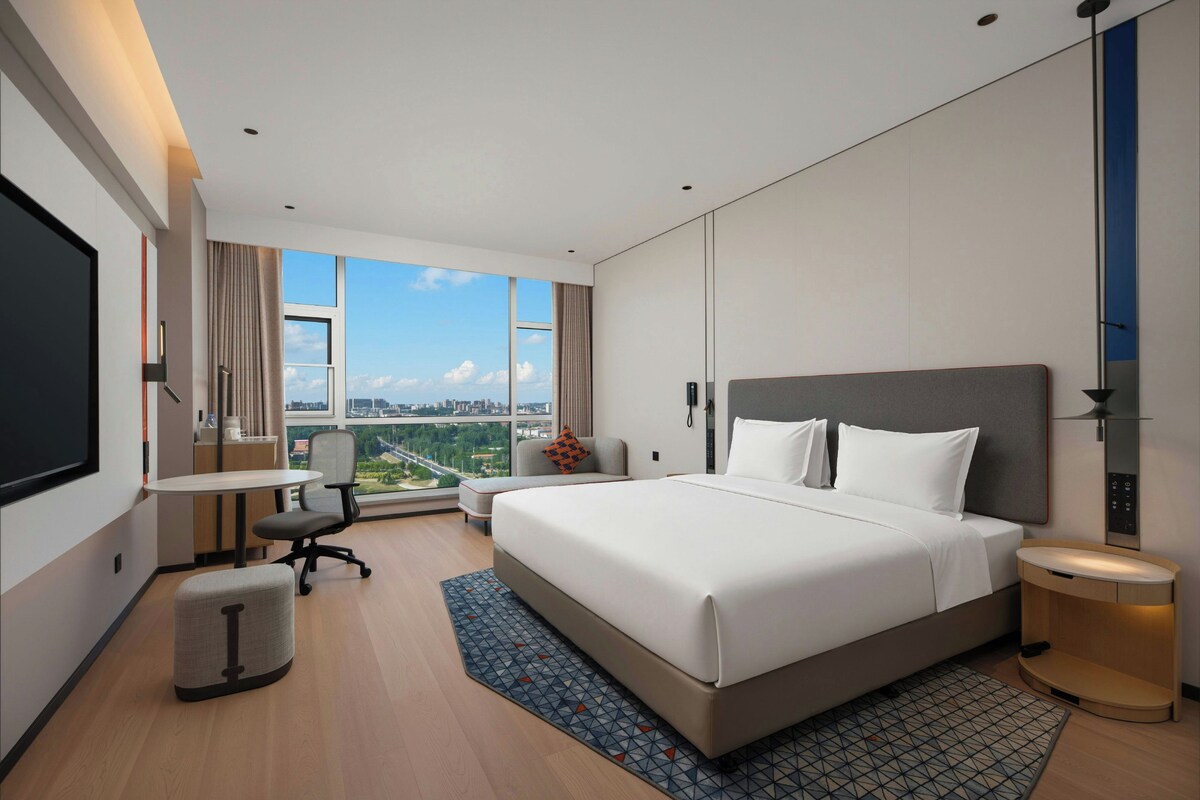 A modern hotel room with a large bed, a desk, a chair, and a view of the city.