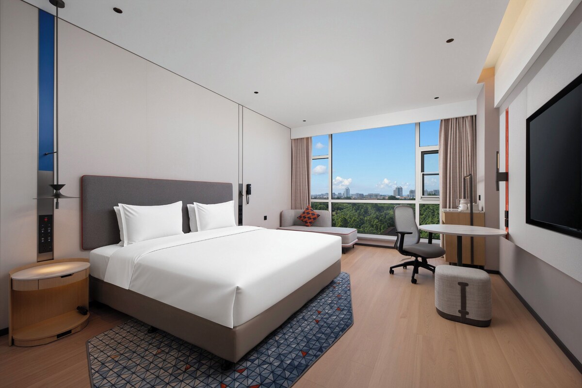 A modern hotel room with a large bed, a desk, a chair, a TV, and a view of the city.