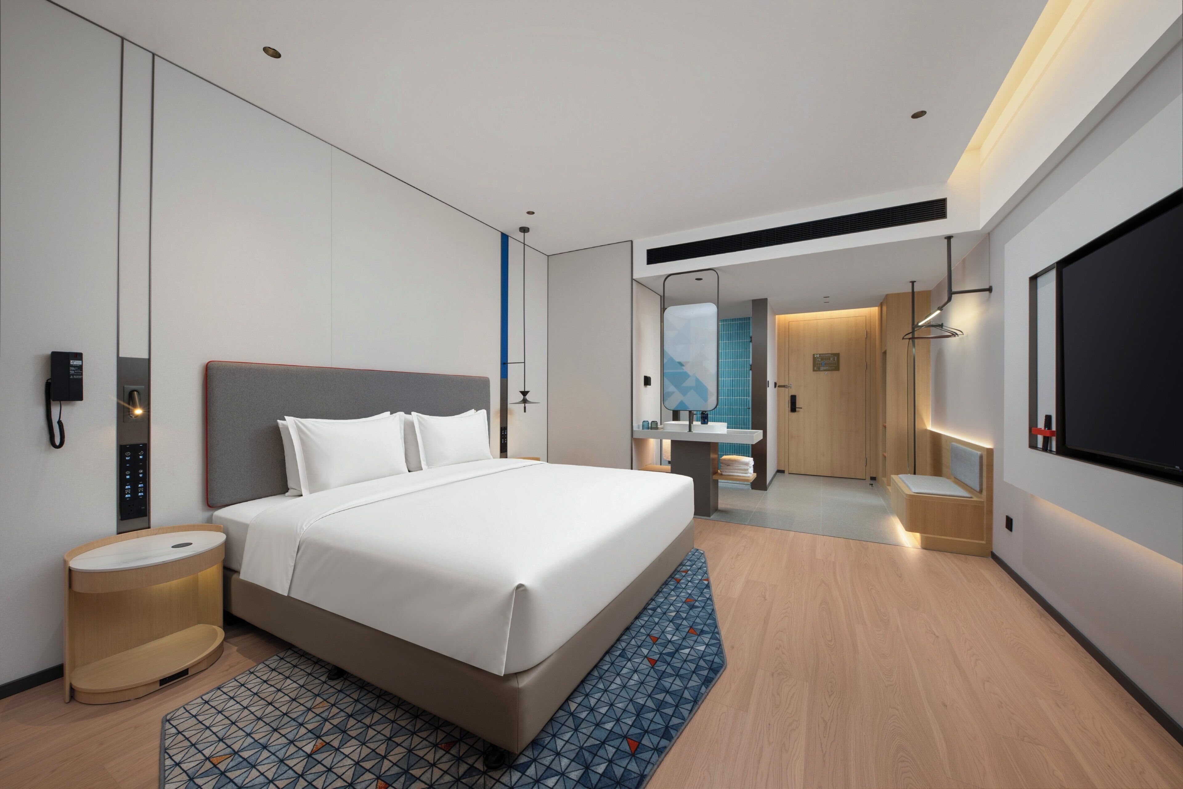A modern hotel room with a large bed, a bedside table, a mirror, a wardrobe, and a bathroom visible through an open door.