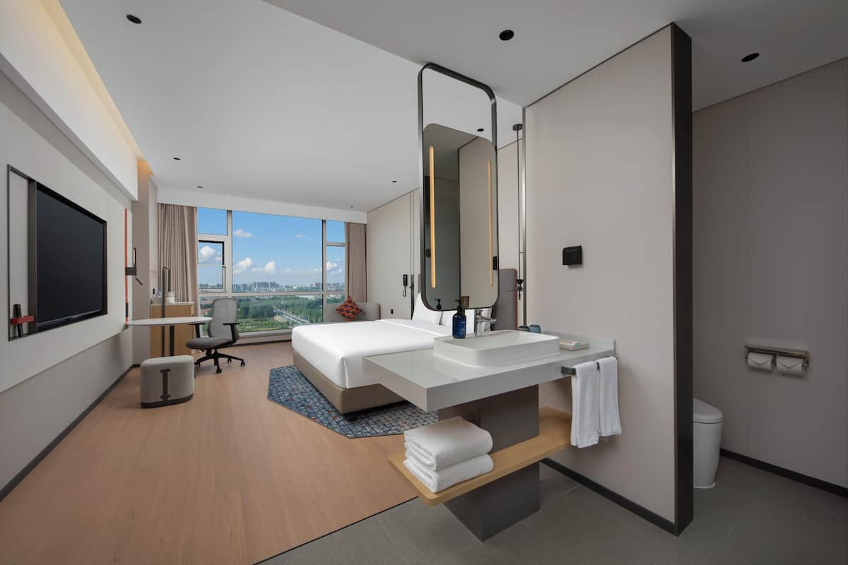 Ett modernt hotellrum med en stor säng, ett skrivbord, ett badrum med spegel och utsikt över staden.