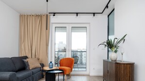 Ein modernes Wohnzimmer mit einer Couch, einem Sessel, einem kleinen Tisch, einem Schrank und einem großen Fenster mit Blick auf Gebäude.