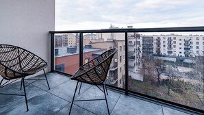 Ein Balkon mit zwei Korbstühlen, einem Kachelboden und Blick auf Gebäude durch ein Glasgeländer.