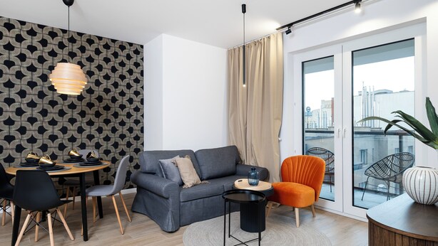 Ein modernes Wohnzimmer mit grauem Sofa, einem Esstisch mit Stühlen, einem kleinen Beistelltisch, einem großen Fenster mit Vorhängen und Blick auf die Stadtlandschaft.