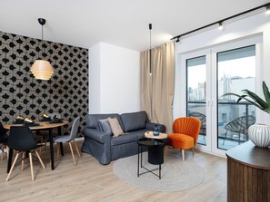 Una sala de estar moderna con un sofá gris, una mesa de comedor con sillas, una pequeña mesita auxiliar, un ventanal con cortinas y vistas a la ciudad.
