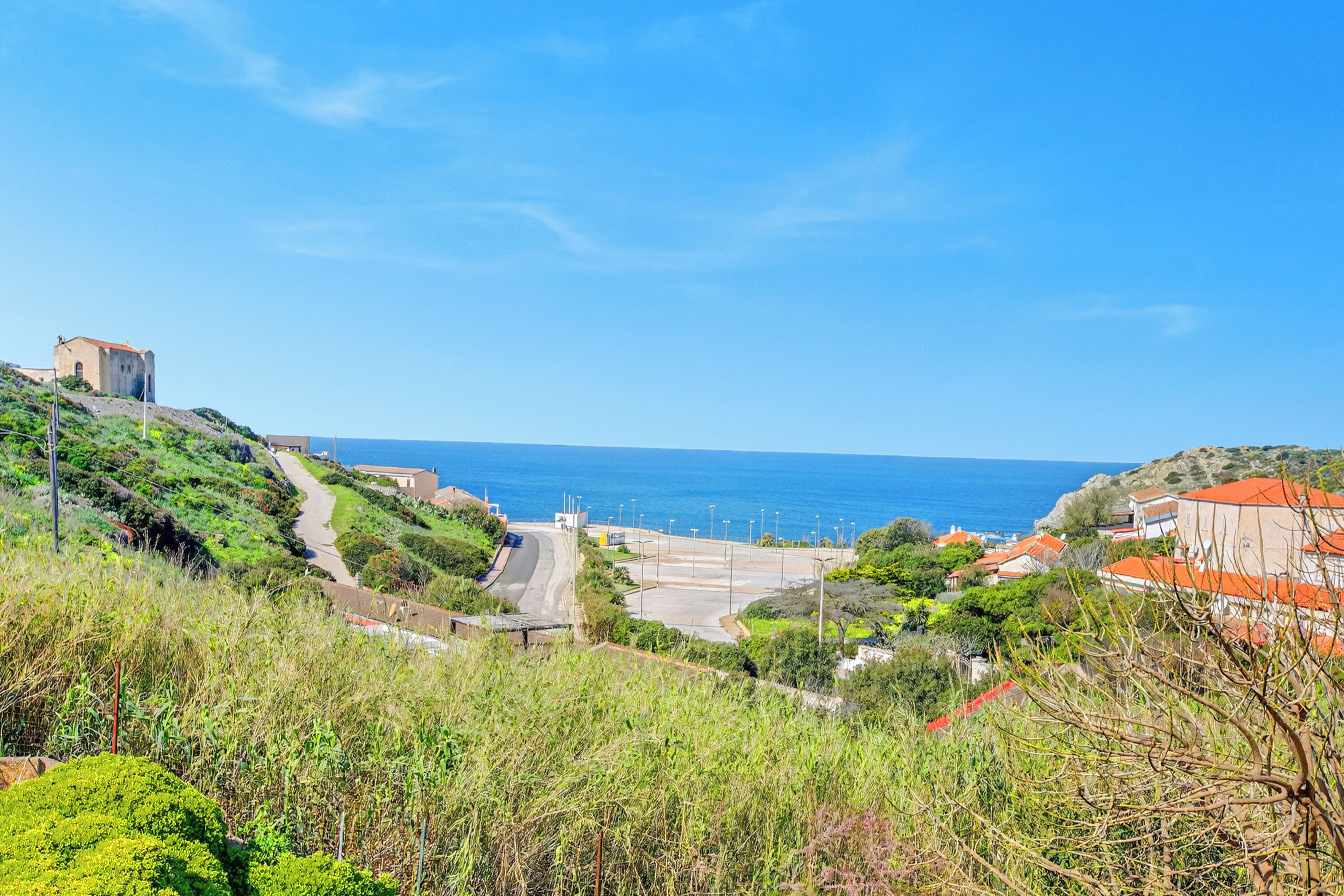 A coastal landscape with a road leading to a building, surrounded by greenery and overlooking the sea.