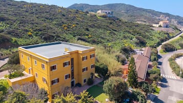 Un edificio giallo a più piani con tetto piatto, circondato da vegetazione e colline sullo sfondo.