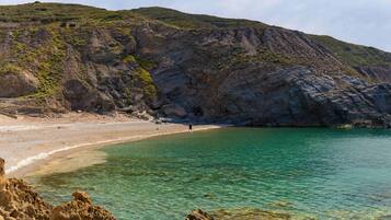 Paesaggio costiero con spiaggia, acqua limpida, scogliere rocciose e una persona che cammina sulla riva.