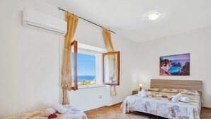 Habitación de hotel con dos camas, una ventana con cortinas, un cuadro de una escena costera y un equipo de aire acondicionado.