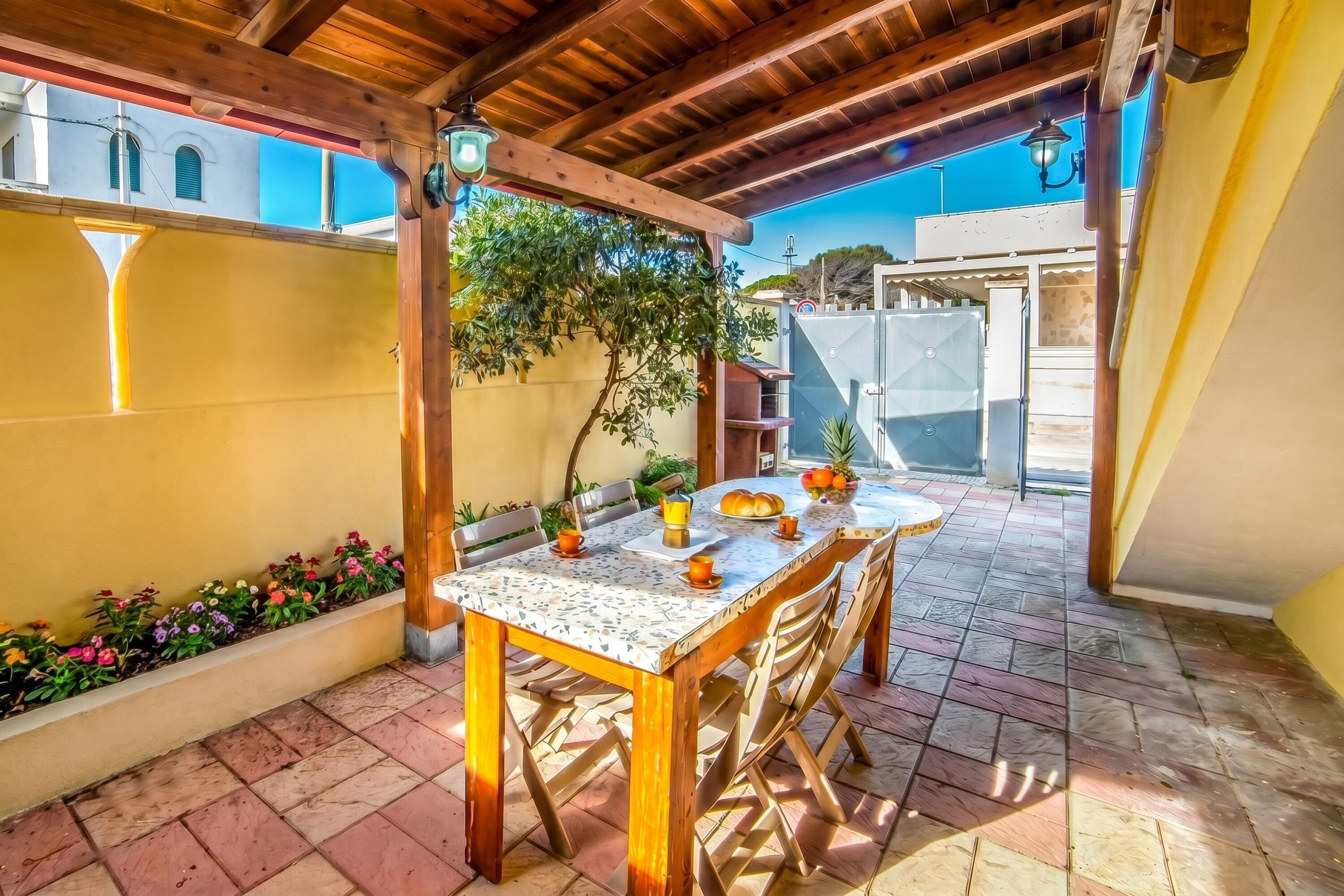 A covered patio with a wooden table and chairs, a tiled floor, and a wall with a window and plants.