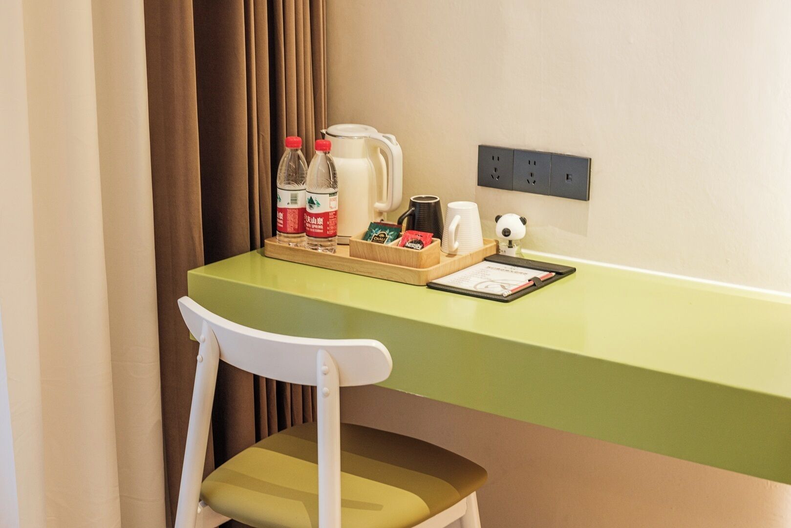 A small desk with a green surface, a white chair, a coffee maker, two bottles of water, a mug, a panda figurine, and a notepad.