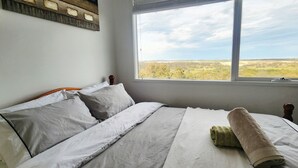 A bedroom with a large window, a bed with white linens, and a view of a landscape.