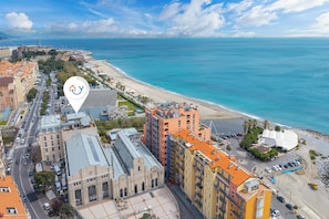 Une vue aérienne d’une ville côtière, avec un mélange de bâtiments modernes et historiques, une plage et un ciel bleu dégagé.