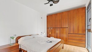Quarto com cama, armário de madeira, mesa de cabeceira, luminária e ventilador de teto.