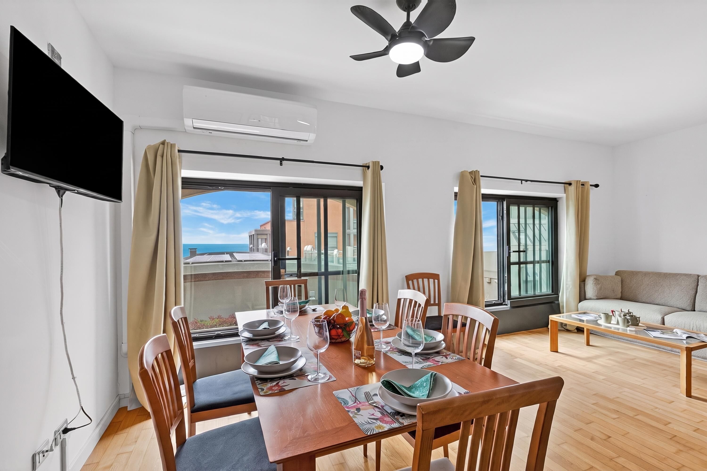 A dining area with a table set for four, chairs, a ceiling fan, and large windows with curtains.