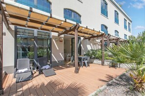 Ein überdachter Außenbereich mit Liegestühlen und einem Tisch, neben einem Gebäude mit großen Fenstern und einer Pergola.