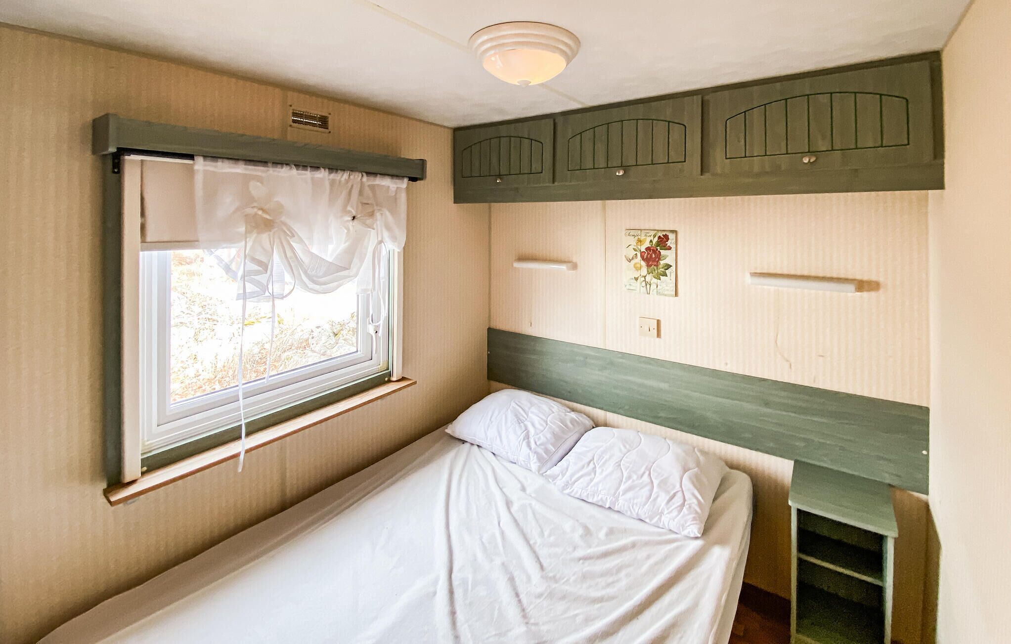 Una cama individual con cabecera, una ventana con cortinas, un estante montado en la pared y una pequeña fotografía.