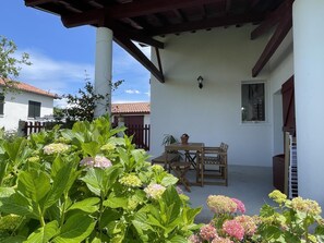 A covered patio with a table and chairs, surrounded by lush greenery and a white building with a red-tiled roof. - HARTZURIENEA - ANHAUX (ANHAUX)
