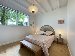 A bedroom with a large window, a bed with a patterned bedspread, a wooden bench, a bedside table with a lamp, and a wall-mounted decorative item. - HARTZURIENEA - ANHAUX (ANHAUX)