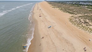 Uma praia extensa com declive suave, alguns guarda-sóis e pessoas caminhando na orla.