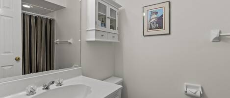 Un baño con lavamanos blanco, espejo, inodoro, armario y un cuadro en la pared.