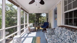 Eine überdachte Veranda mit Deckenventilator, Korbmöbeln, einem gemusterten Sofa und großen Fenstern mit Blick auf Bäume.