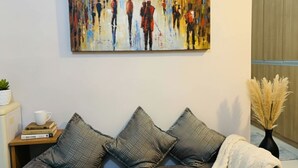 Ein Wohnzimmer mit einer Couch, Kissen, einem Gemälde, einem kleinen Tisch mit Tasse und Büchern sowie einer Vase mit getrockneten Pflanzen.