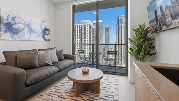 A modern living room with a sofa, a round coffee table, a large window with a city view, and a flat-screen TV mounted on the wall. - 501 First Luxury Ocean View from Balcony Rooftop Pool & GYM 3510 (Miami)
