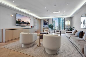 Ein modernes Wohnzimmer mit Fernseher, einer Couch, einem Couchtisch und einem großen Fenster mit Blick auf hohe Gebäude.