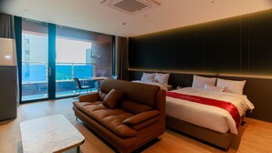 Quarto de hotel com cama, sofá, mesa pequena e vista da cidade por grandes janelas.