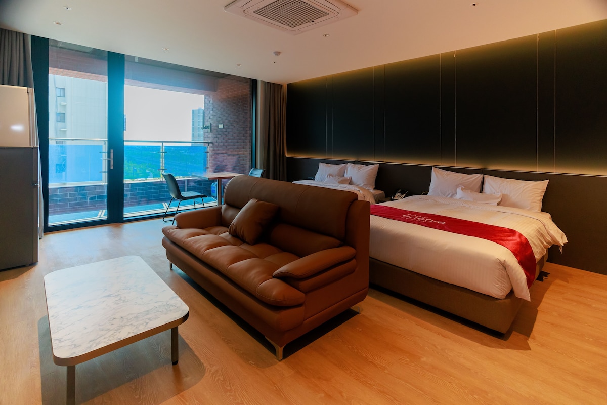 A hotel room with a bed, a sofa, a small table, and a view of the city through large windows.