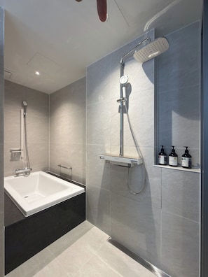 A modern bathroom with a bathtub, a shower with a handheld showerhead, and a shelf with toiletries. - Ramada Encore ASAN (Asan)