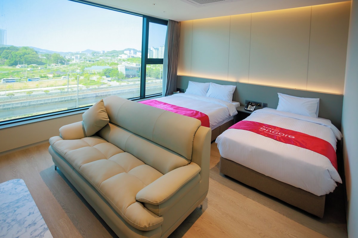 A hotel room with a sofa, two beds, a phone, and a view of the city.