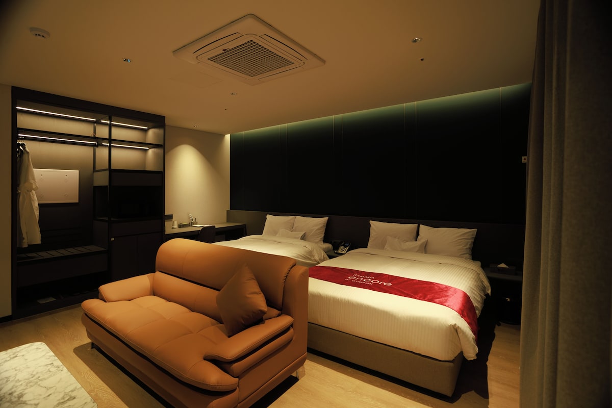 A hotel room with a sofa, a bed with a red bedspread, a bedside table with a phone, and a mirror.