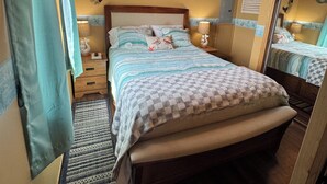 Une chambre avec un plafond en bois, un lit recouvert d’une couverture à carreaux, une table de chevet, une lampe, un miroir fixé au mur et un ventilateur de plafond.