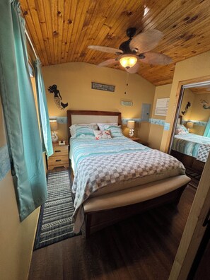 A bedroom with a wooden ceiling, a bed with a checkered bedspread, a nightstand, a lamp, a wall-mounted mirror, and a ceiling fan. - 2 bedroom 1.5 bath - cottage in welcoming Aurora on Cayuga Lake! (Aurora)